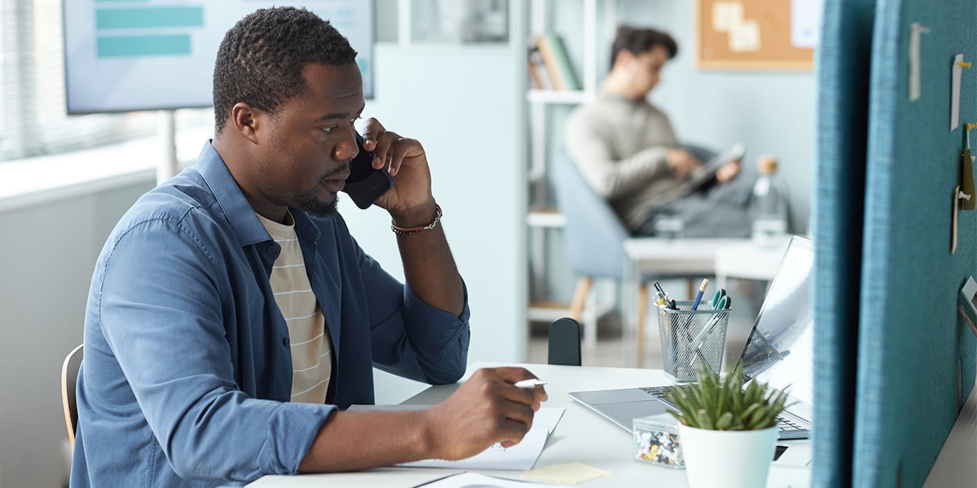 A man on the phone, taking notes at a desk with a laptop, and a plant. A person in background reads quietly.