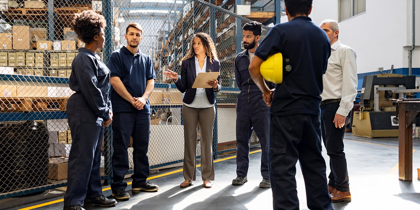 A group of six workers in uniforms discussing in a warehouse setting with boxes in the background.