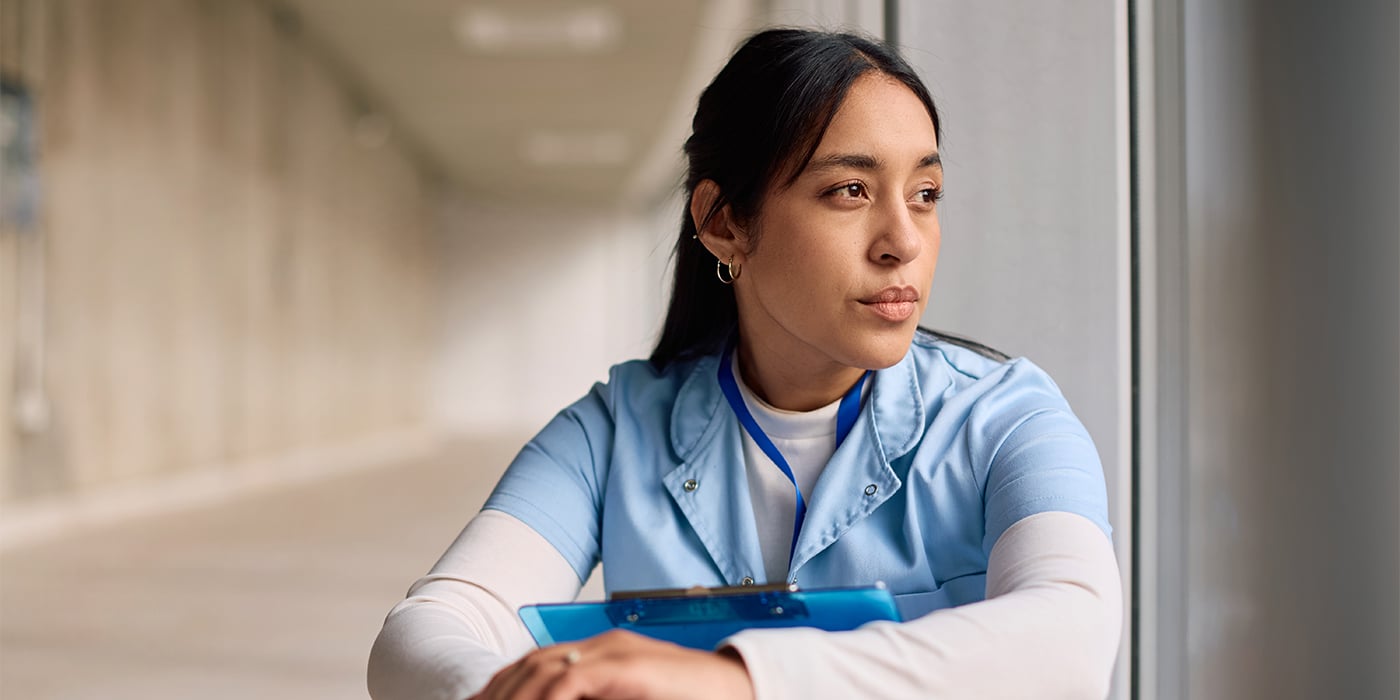 A healthcare worker in a blue uniform leans against a window, looking thoughtful.