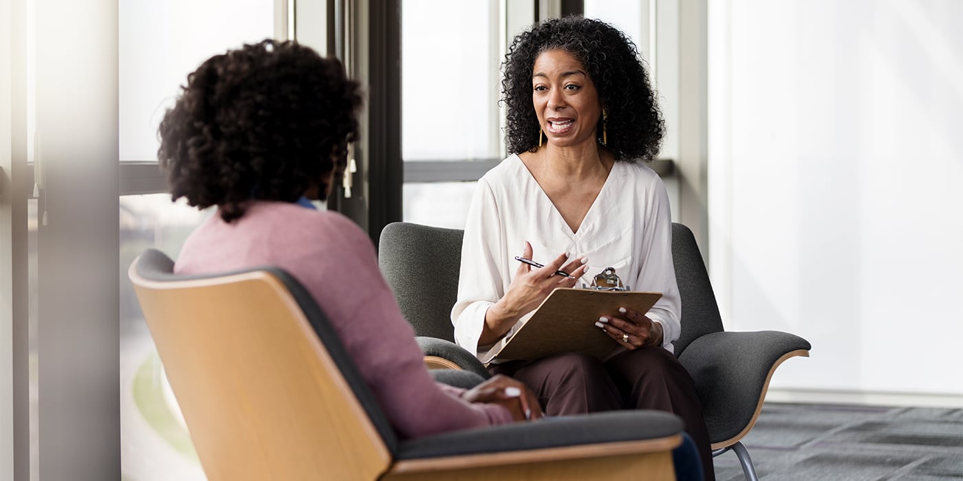 A woman with curly hair actively listening and taking notes on a clipboard while talking to another woman.
