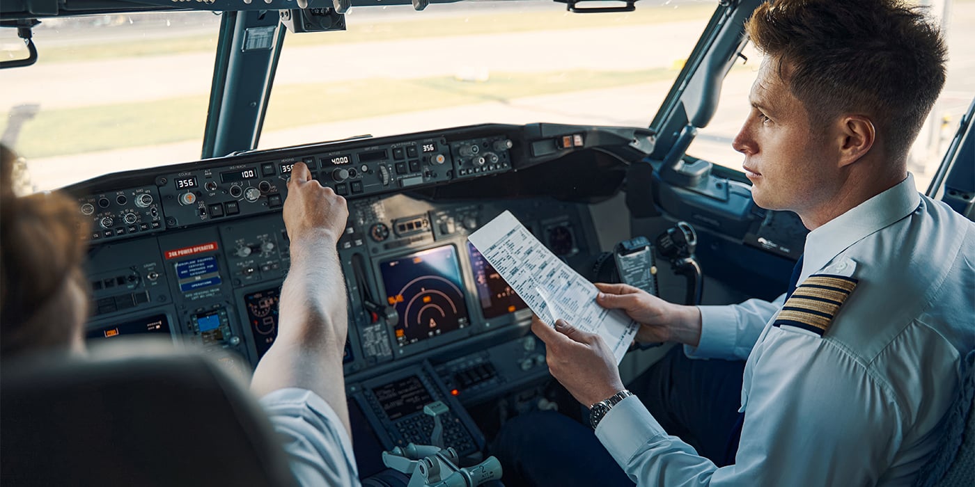 Two pilots in an airplane cockpit, one adjusting controls and the other checking a flight manual.
