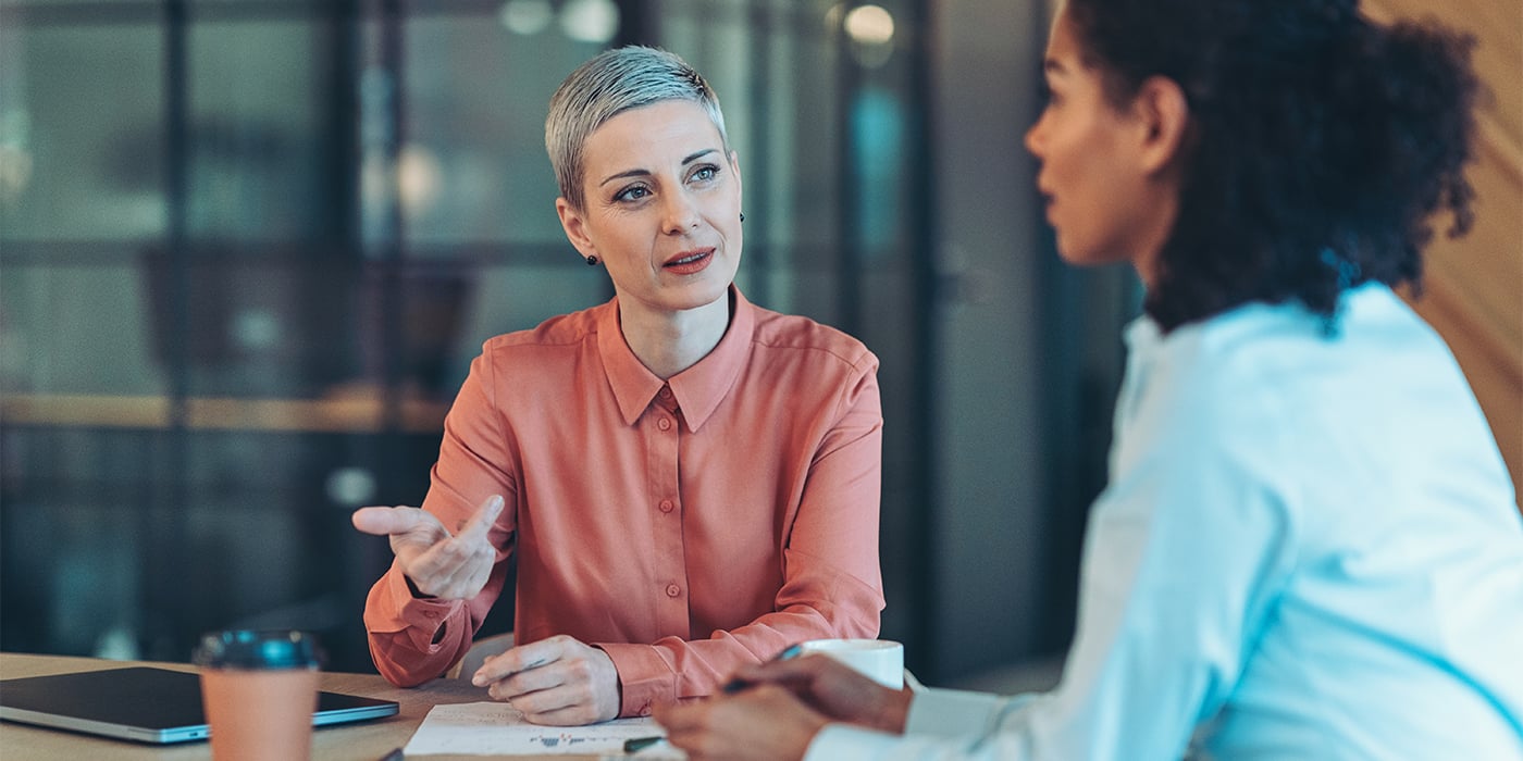 Two women in a meeting, one in a pink shirt speaking while the other listens attentively.
